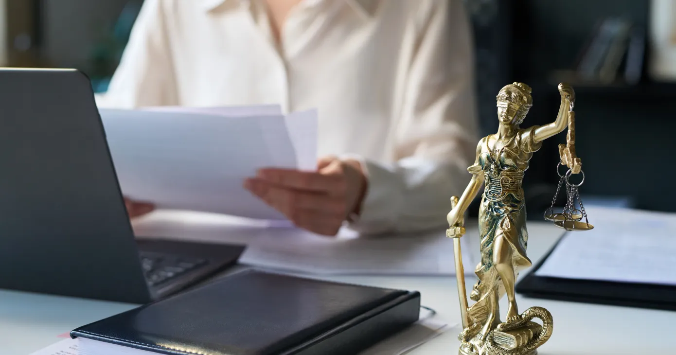 Bronze Lady Justice statuette on desk of attorney in law firm, holding scales and sword, and female lawyer working with documents in blurred background